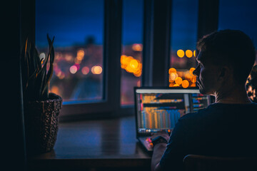 Wall Mural - a young man programmer coding on a laptop in the dark with a view of the lights of the night city, c