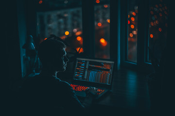 Wall Mural - a young man programmer coding on a laptop in the dark with a view of the lights of the night city, c