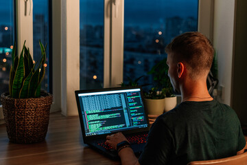 Wall Mural - a young male programmer in front of a window with a lights of night city, on his laptop screen is co