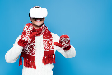 Smiling man wearing vr headset, touching with hands in mittens isolated on blue