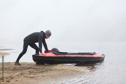 Fotografía Man pulling canoe into water, wearing black clothing, posing on bank of river, handsome sports man doing water sport in foggy autumn day, canoeing in cloudy morning