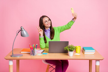 Wall Mural - Photo of positive high school girl sit table make v-sign selfie smartphone wear green shirt isolated pink color background