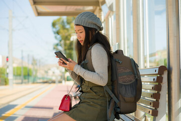 Canvas Print - outdoors lifestyle portrait of young beautiful and happy Asian Korean woman waiting the train sitting on station platform bench using mobile phone checking schedule online