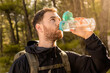 Portrait of young male hiker drinking water from his transparent bottle with background sunlight in a mountain forest with warm colors and lots of light.