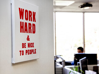A sign in an office that states Work Hard And Be Nice To People in red letters. An office worker is in the background