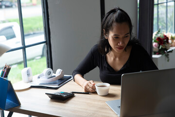 Wall Mural - Remote working at Home office Concept. Businesswoman working online via laptop and drink coffee. Asian young entrepreneur watching webinars and talking during meeting video conferences calls with team