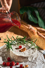 Camembert on parchment paper, close-up on a wooden board. Cranberry sauce is poured over cheese from a jug. On cheese rosemary and walnuts, next to an hour with cranberries. Dark background, sunlight