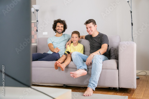 Happy gay fathers and son watching funny TV show at home, sitting on couch in living room, smiling and laughing. Family and home entertainment concept