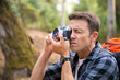 © Mangostar - Focused man shooting landshate and walking in forest. Caucasian traveler exploring nature, holding camera, taking photo and carrying backpack. Tourism, adventure and summer vacation concept