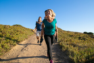 Cheerful family couple and children enjoying hiking in countryside, walking on path. Two kids riding on parents back and neck. Front view. Nature and recreation concept