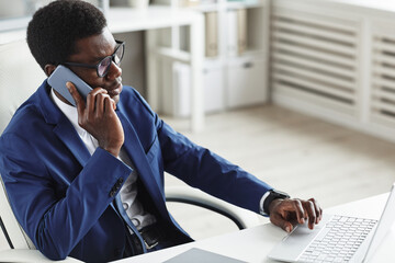 Wall Mural - African businessman in suit sitting at his workplace typing on computer keyboard and talking on mobile phone at office