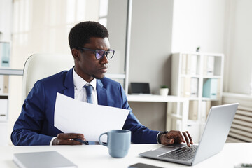 Wall Mural - Young African businessman sitting at the table in front of laptop he typing documents and doing paperwork at office