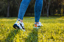 White Sneakers With Black Inserts On Female Legs Dressed In Jeans On Green Grass