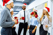 © Shopping King Louie - Happy working team in Santa Claus hats, during a break in the office, greets the New Year and drinks a drink from red mugs