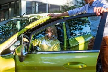 A woman with blonde hair is sitting in a yellow car and a man in a shirt is standing near. Auto business, car sale, consumerism and people concept - happy woman with car dealer. 