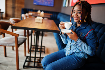 Wall Mural - Lovely african american woman with dreadlocks in blue stylish jeans jacket at cafe. Beautiful cool fashionable black young girl indoor drink coffee.