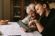 Smiling caucasian grandmother and beautiful granddaughter in the kitchen looking at the tablet. The granddaughter teaches her grandmother to use new technology