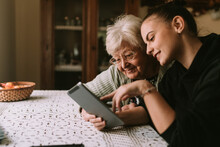 Smiling Caucasian Grandmother And Beautiful Granddaughter In The Kitchen Looking At The Tablet. The Granddaughter Teaches Her Grandmother To Use New Technology