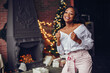 © prostooleh - Beautiful girl in a decorated room. Woman near Christmas tree. Black lady in a white blouse