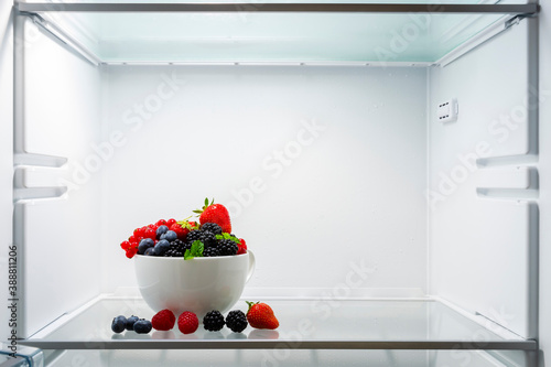 Mix of ripe strawberries, blueberries, raspberries, blackberries, and red currants in a white bowl and on the glass shelf in a fridge. Mix of berries reach of vitamins for healthy eating.