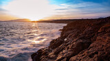 Fototapeta Desenie - Ocean landscape, rocks next to the sea and the waves crashing in the red rocks. Sunshine in the sunset with blue sky and the sun reflections on the sea.