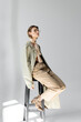 © LIGHTFIELD STUDIOS - full length of young woman in glasses, trench coat and scarf sitting on stool while posing on grey