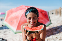 Close-up Of Smiling Woman Holding Watermelon While Sitting Against Red Umbrella At Beach