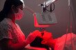 © Space_Cat - Young woman having red LED light facial therapy treatment in beauty salon. Beautician wearing face mask maintaining safety procedures during appointment. Beauty, new normal and wellness