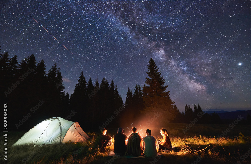 Evening summer camping, spruce forest on background, sky with falling stars and milky way. Group of five friends sitting together around campfire in mountains, enjoying fresh air near illuminated tent - obrazy, fototapety, plakaty 