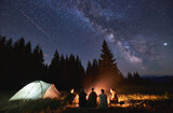 Fototapeta  - Evening summer camping, spruce forest on background, sky with falling stars and milky way. Group of five friends sitting together around campfire in mountains, enjoying fresh air near illuminated tent