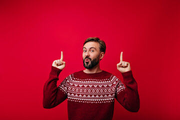 Enthusiastic white man posing with smile on red background. Joyful bearded guy in scandinavian sweater having fun.