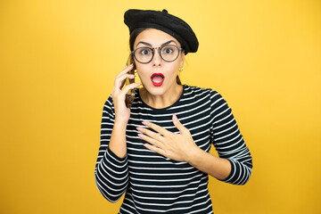 Wall Mural - Young beautiful brunette woman wearing french beret and glasses over yellow background talking on the phone with a worried expression