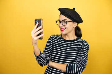 Wall Mural - Young beautiful brunette woman wearing french beret and glasses over yellow background taking a selfie with her phone