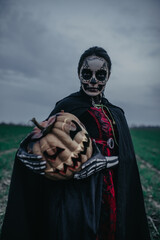 Wall Mural - Woman stands among field in Halloween costume of death and jack-o-lantern against background dark sky.