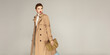© Beauty Agent Studio - Autumn clothing concept. Fashionable young model posing against grey background dressed in trench coat and holding stylish  bag.