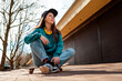 © _KUBE_ - A young beautiful Caucasian woman sits relaxed on a skateboard and looks away. In the background, a street with trees. Bottom view. Concept of sports lifestyle and street culture