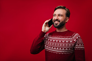 Carefree young man talking on phone on red background. Cute male model in new year sweater calling someone.