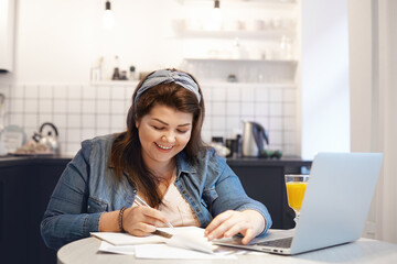 Education, modern technology, occupation and communication concept. Chubby happy female student smiling joyfully while doing homework, recollecting something funny, sitting in front of open laptop