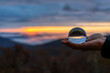 Hand holding crystal glass lens ball view of round globe with reflection of fog mist clouds over blue ridge mountains at colorful sunrise in Wintergreen, Virginia