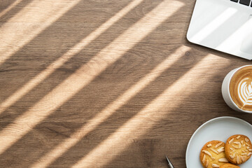 Wood office desk table with dish of cookies, cup of latte coffee and supplies. Top view with copy space, flat lay. Working from home in the morning concept.