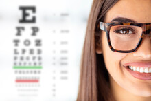 Close Up Of Young Woman Wearing Eyeglasses With Eyechart In The Background. Eyewear. Closeup View Of Young Woman And Blurred Eye Chart On Background. Space For Text