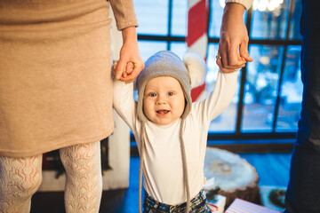 Funny little baby boy 1 year old learning walk home in winter in a decorated New Year house. Young family dad and mom hold by the hands of his son in the loft interior wooden floor near the window