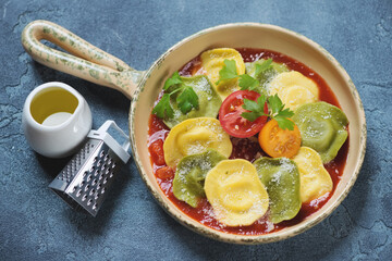 Beige serving pan with italian ravioli on tomato sauce bedding, horizontal shot on a blue stone background, elevated view