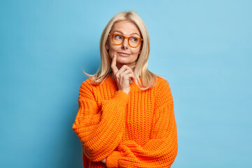 Beautiful middle aged wrinkled woman keeps index finger on cheek stands in thoughtful pose daydreams about something wears knitted sweater isolated over blue background. Need to think deeply