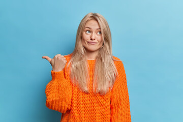 Wall Mural - Uninterested blonde woman dressed in knitted jumper points away and shows direction dissatisfied with something poses against blue background. You may go there. What about visiting this place