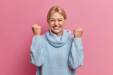 Cheerful successful blonde woman celebrates victory raises hands with clenched fists smiles toothily wears blue sweater isolated over pink background. Overjoyed young female receives great news