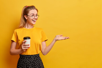 Sincere positive girl drinks aromatic coffee from disposable cup keeps hand raised over yellow empty background happy to see friend far away wears yellow t shirt and skirt being in good mood