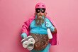 Stunned bearded man going to clean house stares bugged eyes has thick beard with clothespins holds plunger wrapped by toilet paper and cleaning detergent dressed in superhero costume isolated on pink