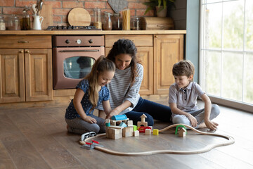 Loving young Caucasian mother have fun engaged in funny game with two little children at home kitchen. Happy caring mom and smiling small kids play together with wooden blocks toys on floor.