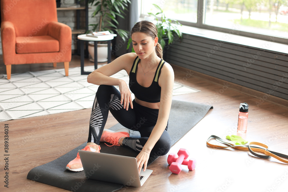 Beautiful slim sporty woman in sportswear is sitting on the floor with dumbbells and bottle of water and is using a laptop at home in the living room. Healthy lifestyle. Stay at home activities. - obrazy, fototapety, plakaty 